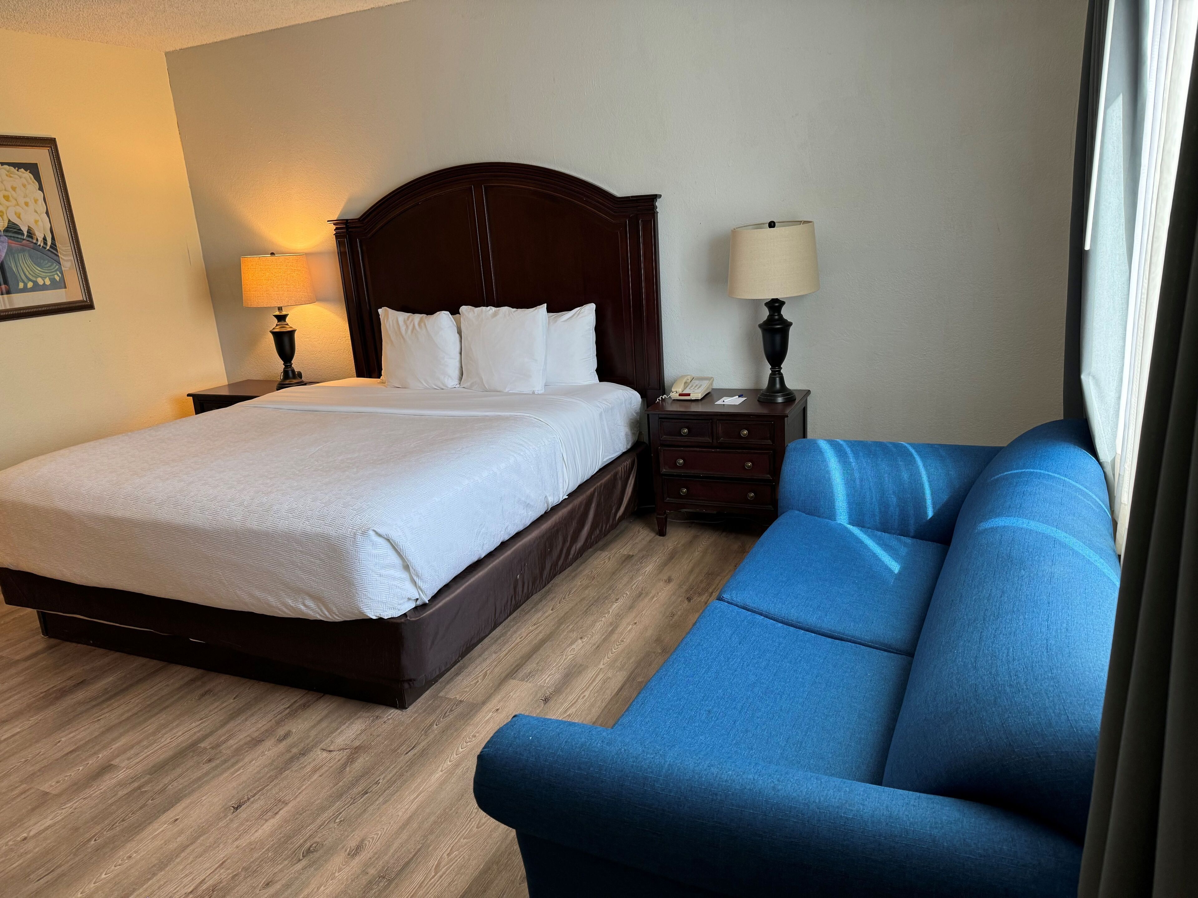 Een hotelkamer met een bed, een nachtkastje, een lamp, een blauwe bank en een schilderij aan de muur.