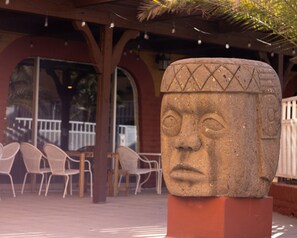 Escultura de una cabeza de piedra en un entorno exterior con una zona cubierta y sillas.