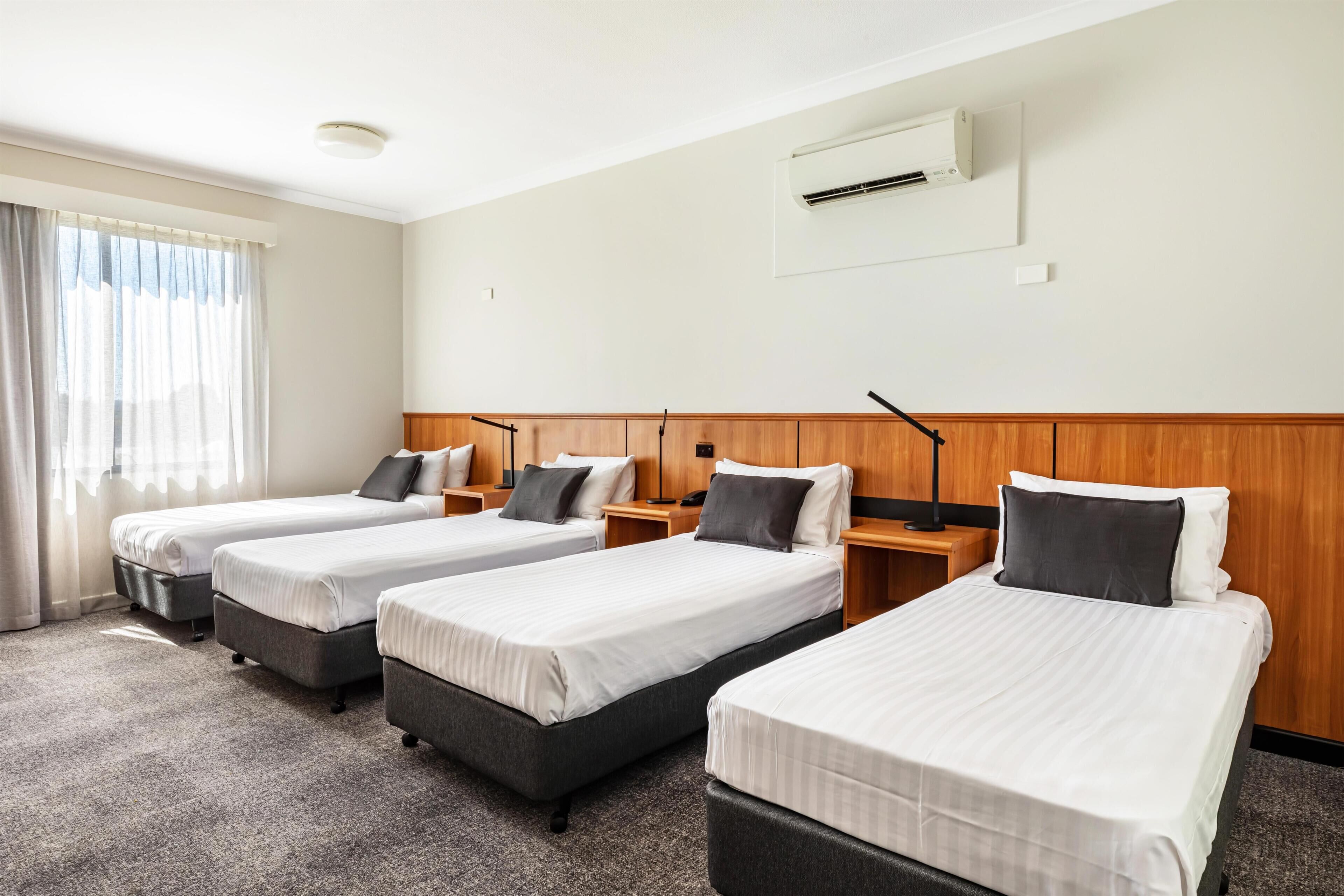 A hotel room with three single beds, a wooden headboard, a window with curtains, an air conditioning unit, and a carpeted floor.