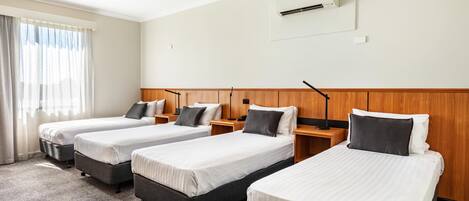 Una habitación de hotel con tres camas individuales, cabecero de madera, ventana con cortinas, unidad de aire acondicionado y suelo alfombrado.