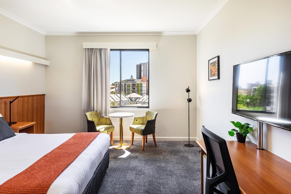 A hotel room with a bed, a desk, a chair, a small table, a potted plant, a TV, and a window with a city view.