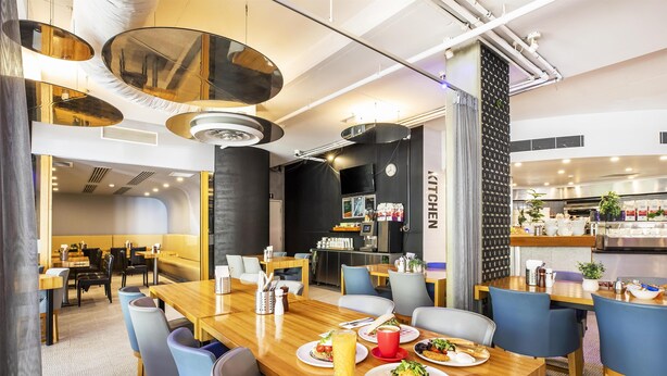 A modern dining area with wooden tables and chairs, a kitchen in the background, and a clock on the wall. - Metro Hotel Perth City (East Perth)