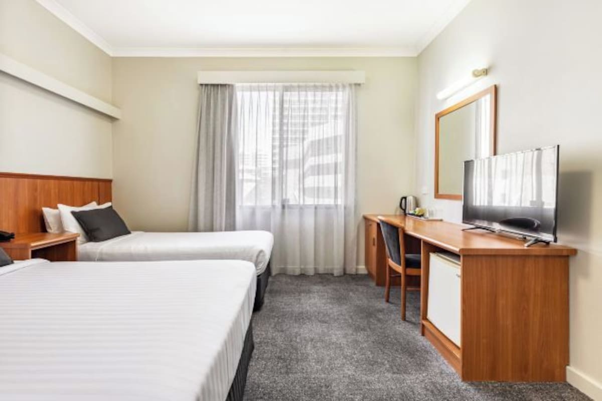 Habitación de hotel con dos camas, un escritorio con televisor, una silla y una ventana con cortinas.
