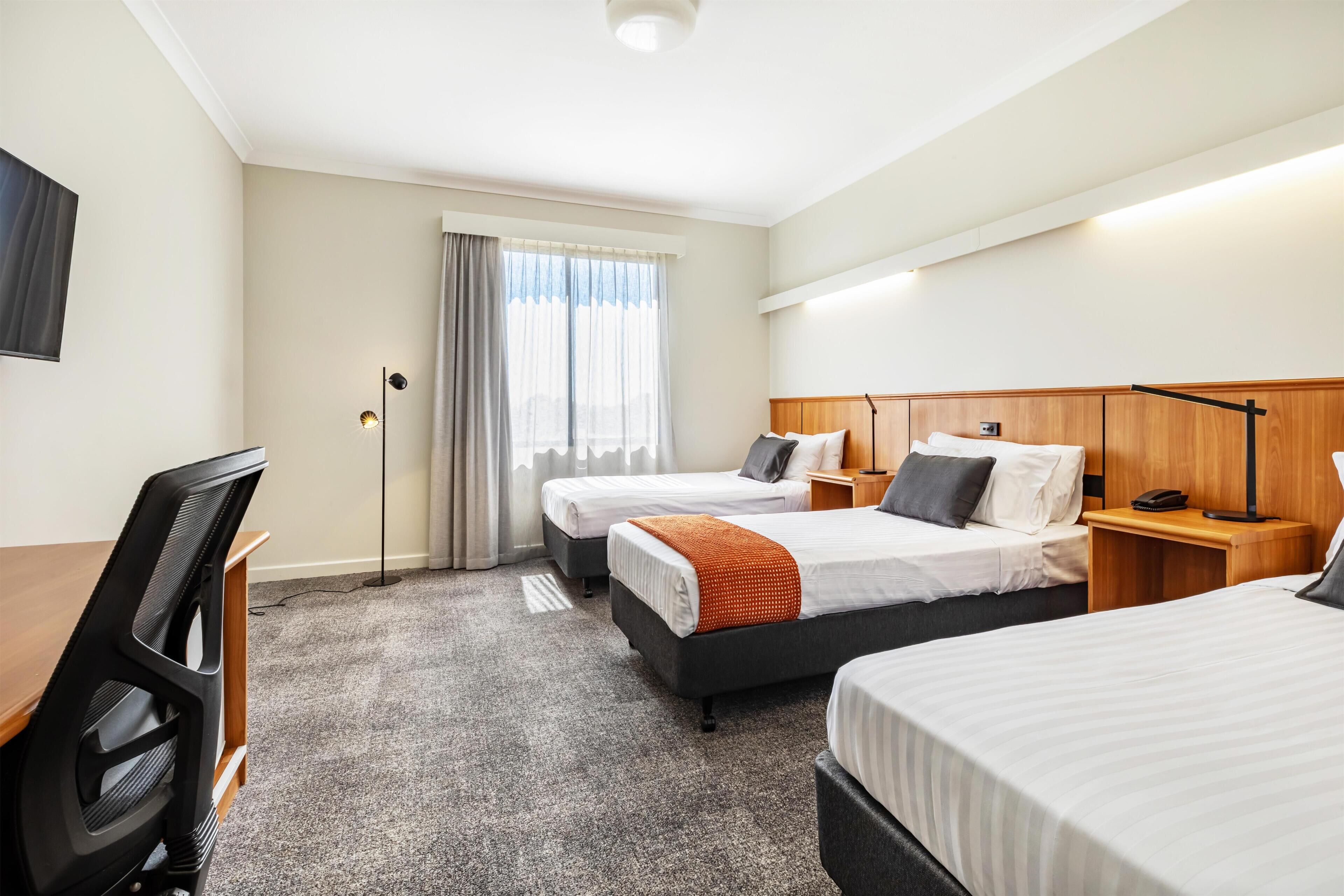 Quarto de hotel com duas camas, uma mesa, uma cadeira, um abajur e uma janela com cortinas.