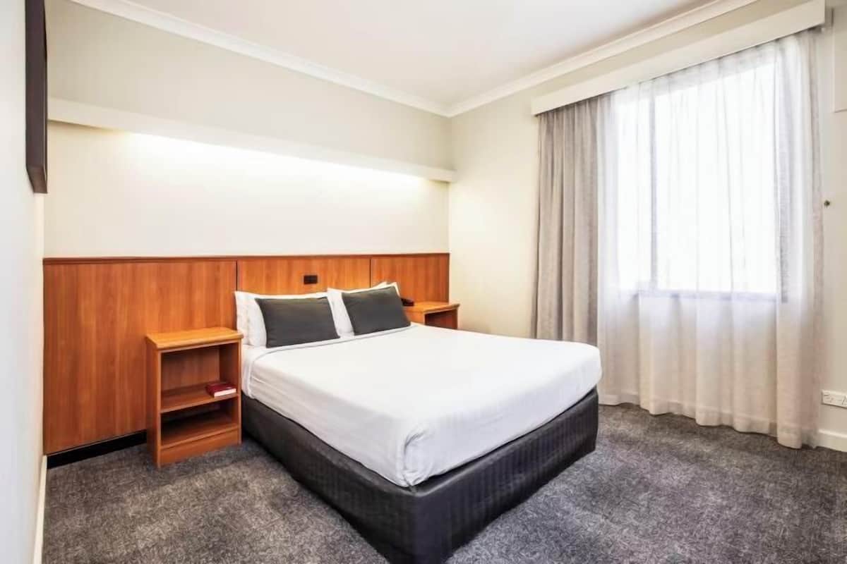A hotel room with a bed, wooden headboard, two bedside tables, a nightstand, and a window with curtains.