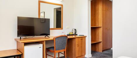 A compact hotel room with a wooden desk, a small refrigerator, a television, a chair, and a wardrobe.