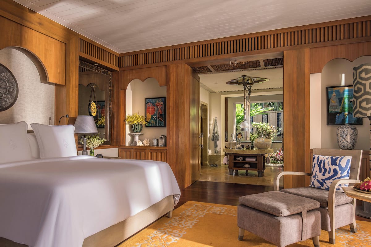 Un dormitorio espacioso con una cama grande, una zona de estar con una silla y una mesa, paneles de madera y vistas a un jardín exuberante.