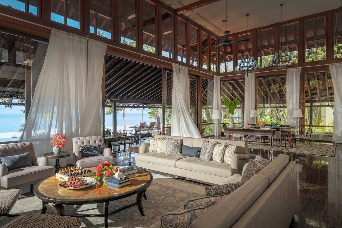 Amplia sala de estar con un sofá grande, una mesa de centro con objetos decorativos y vistas al exterior a través de amplias ventanas.