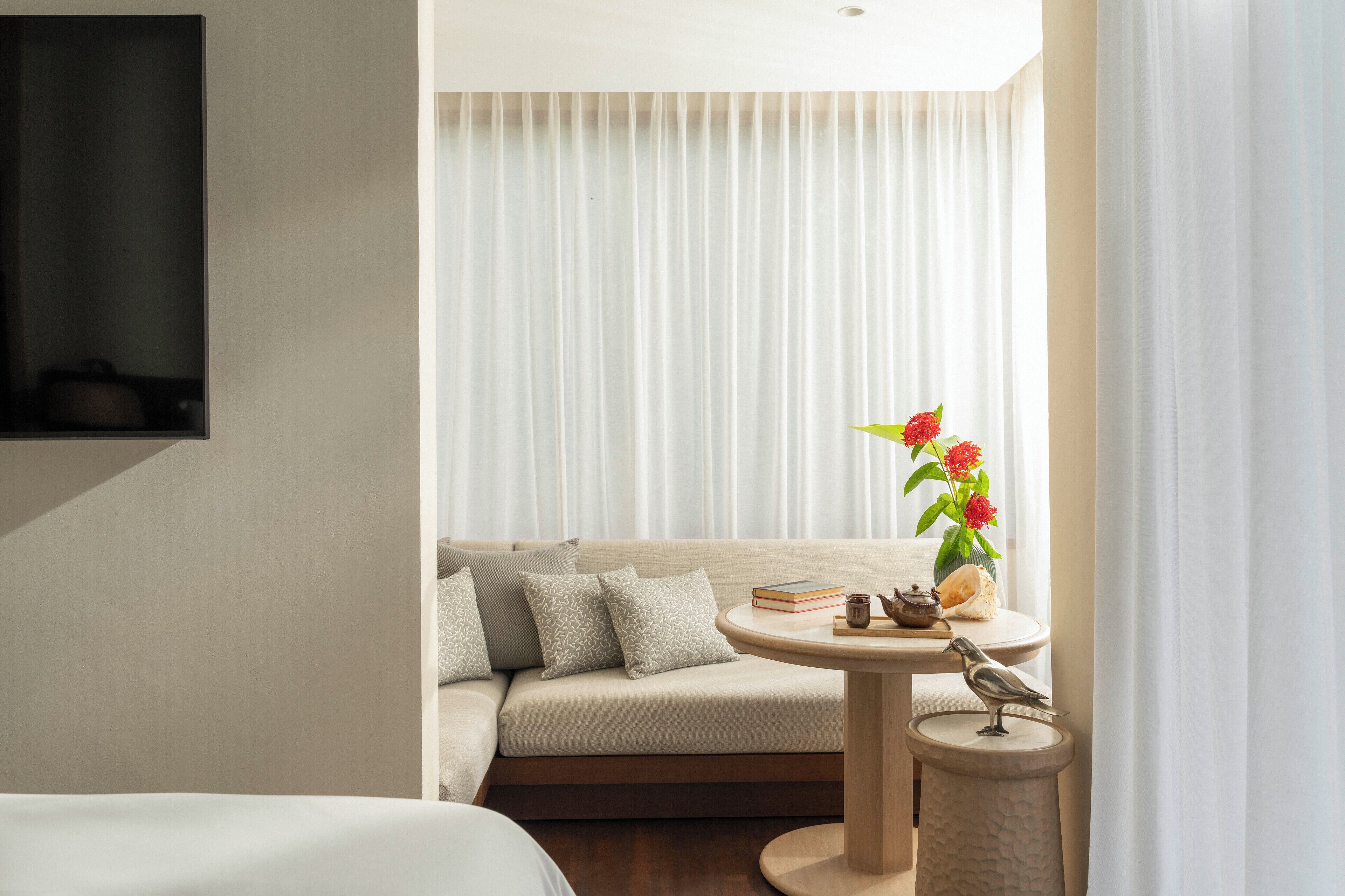 Une chambre d’hôtel moderne avec un canapé, une petite table ronde ornée d’un vase de fleurs, un oiseau décoratif et une bibliothèque.
