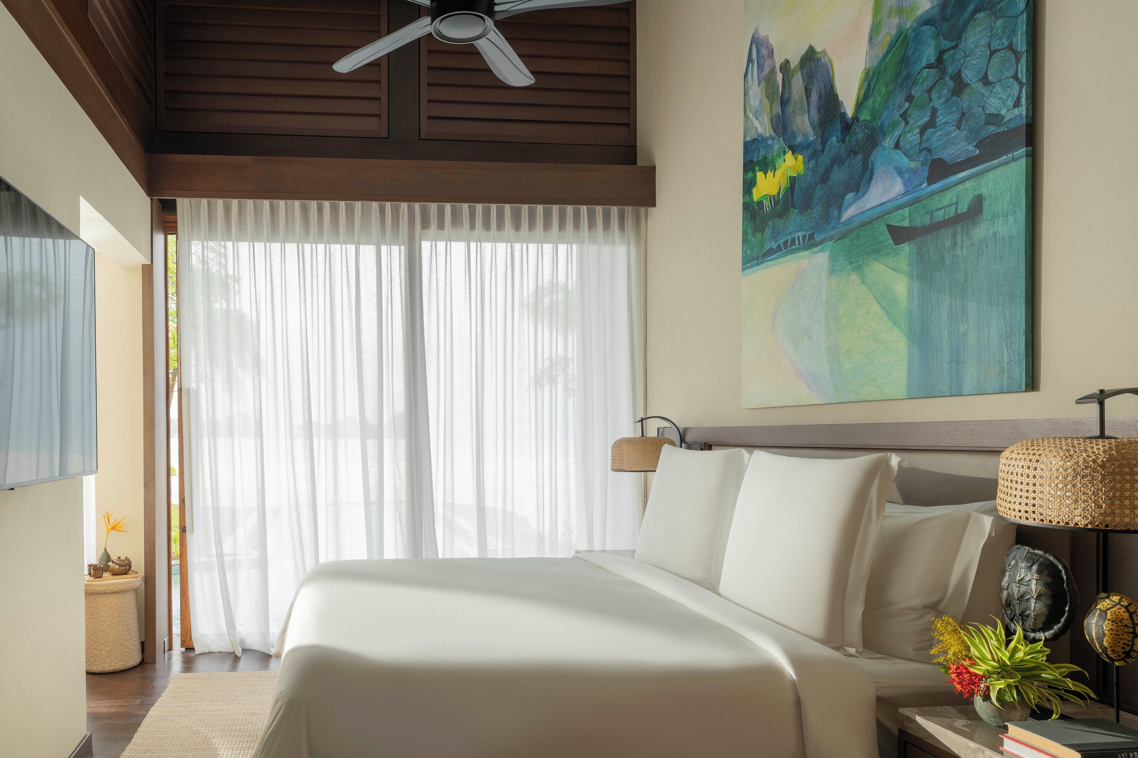 A hotel room with a large bed, a ceiling fan, a painting on the wall, and a window with sheer curtains.