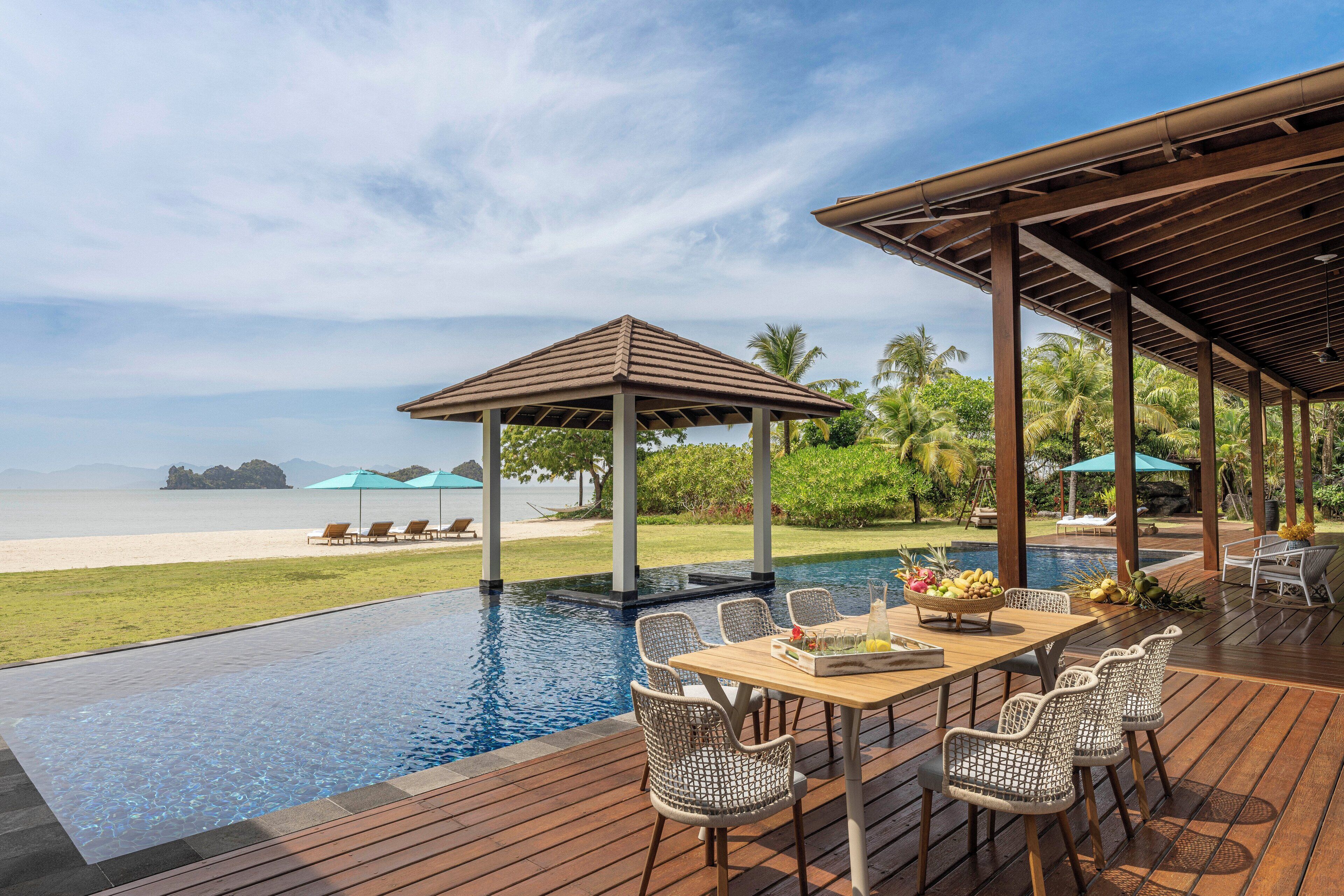 Une piscine à débordement avec une terrasse en bois, des chaises longues et un espace abrité offrant une vue sur la plage et l’océan.