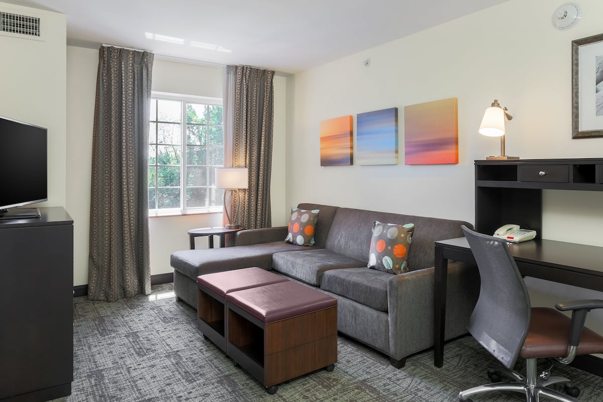 A modern hotel room with a sofa, a desk, a chair, a TV, and artwork on the wall.