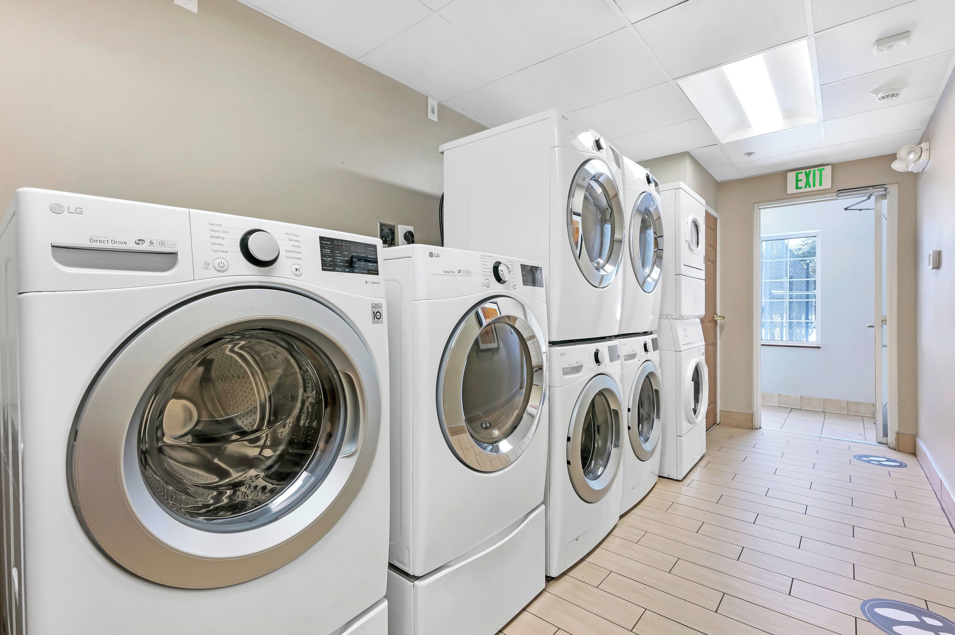 A laundry facility with multiple white washing machines and dryers, an exit sign, and a tiled floor.