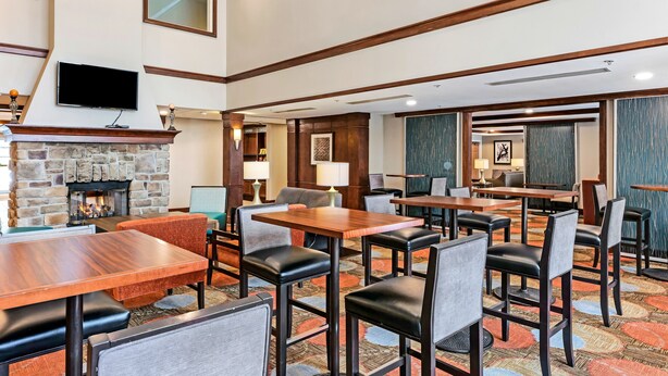A hotel lobby with a stone fireplace, wooden tables and chairs, a flat-screen TV mounted above the fireplace, and a patterned carpet. - Staybridge Suites Philadelphia-Mt. Laurel by IHG (Mount Laurel)