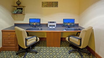 Un salón de conferencias con dos escritorios, cada uno con una computadora, teclado y ratón. Hay dos sillas, una impresora y un estante decorativo con una planta.