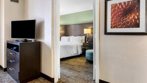 A hotel room with a flat-screen TV on a dresser, a bed with white linens, a nightstand with a lamp, and a framed floral artwork on the wall. - Staybridge Suites Philadelphia-Mt. Laurel by IHG (Mount Laurel)