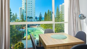 A room with a large window, a dining table with chairs, and a view of tall buildings and greenery. - Maldives Resort (Main Beach)