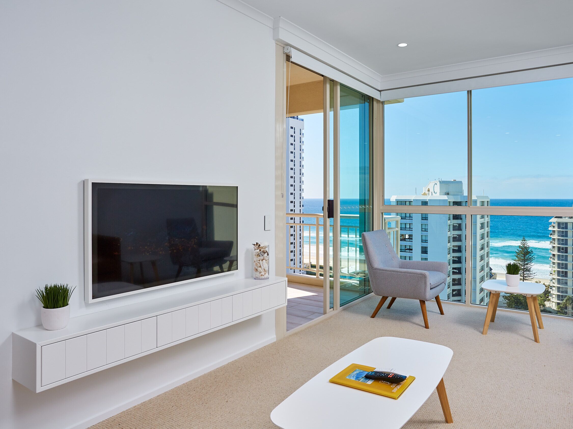 Una sala de estar moderna con un televisor de pantalla plana grande, un área de estar confortable y un balcón con vistas al océano y edificios.