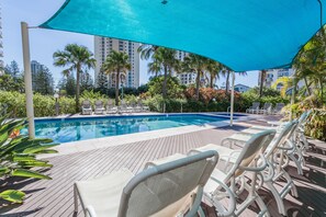 A poolside area with lounge chairs and a large umbrella, surrounded by palm trees and buildings. - Maldives Resort (Main Beach)