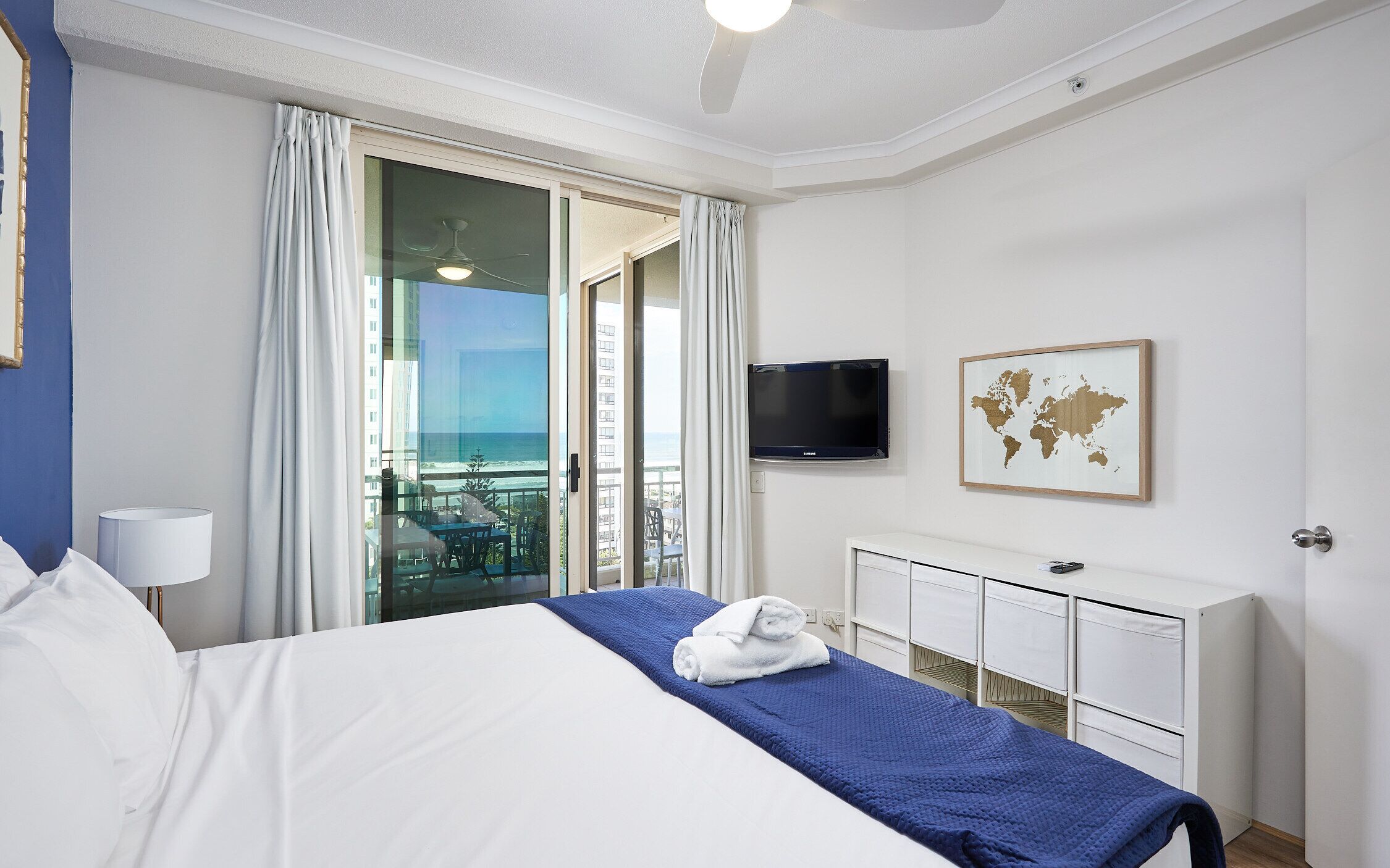 Habitación de hotel con cama, televisión, un cuadro de mapa mundial y balcón con vistas al océano.