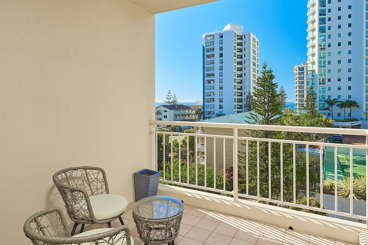 A balcony with wicker chairs and a small table, overlooking tall buildings and a clear blue sky.