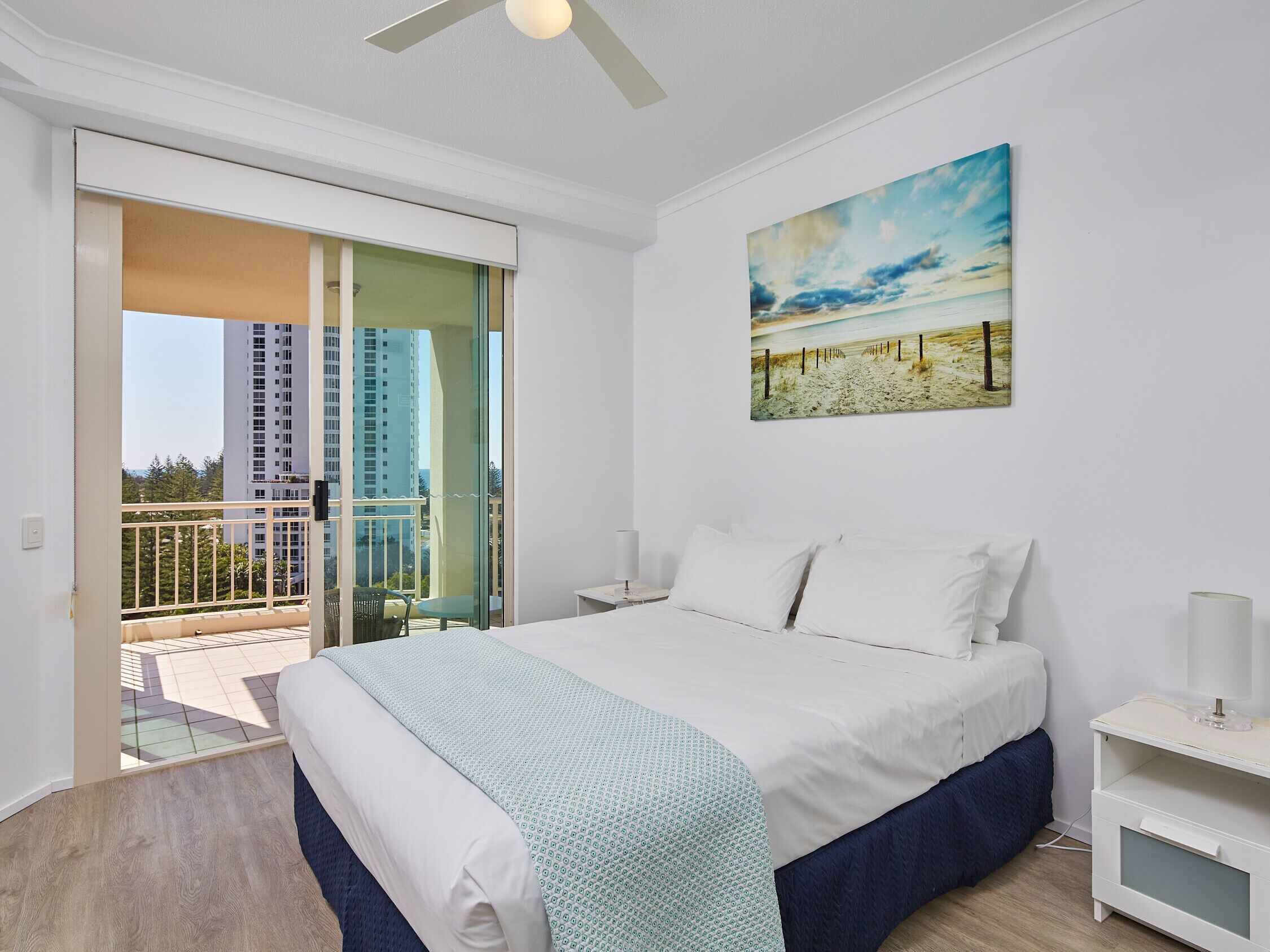Un dormitorio con cama, mesita de noche, un cuadro y un balcón con vistas a edificios.