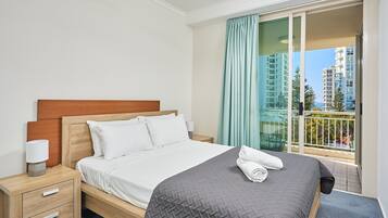 Een hotelkamer met een bed, nachtkastje, lamp en uitzicht op een balkon met gebouwen.