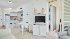 A modern kitchen with white cabinets, a dining area with a white table and blue stools, a TV on a white cabinet, and a grey sofa in the living area. - Maldives Resort (Main Beach)