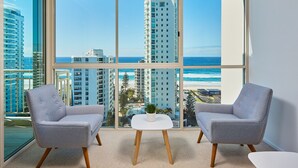 A modern room with large windows, two grey upholstered chairs, a small white table, and a view of the ocean and high-rise buildings. - Maldives Resort (Main Beach)
