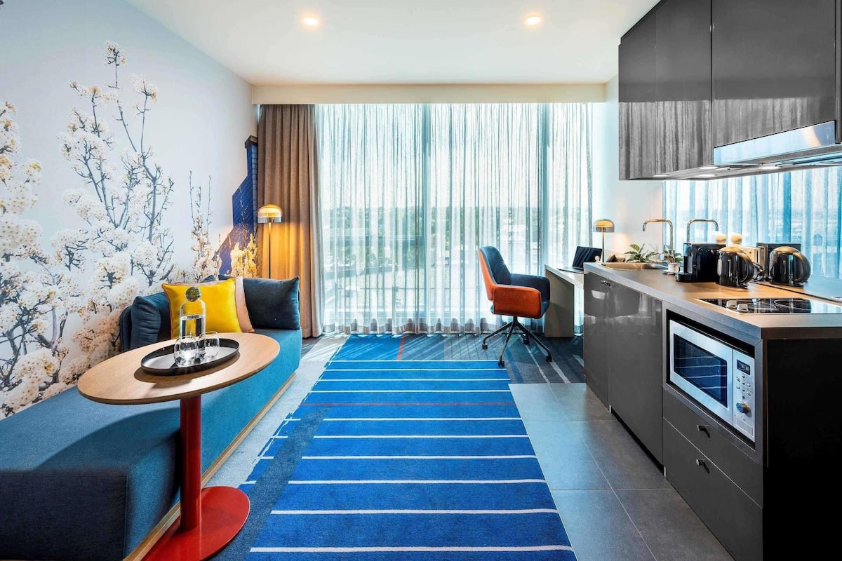 Una habitación de hotel moderna con sofá, una pequeña mesa redonda, una zona de cocina con estufa y un amplio ventanal con cortinas.