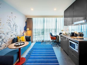 Una habitación de hotel moderna con sofá, una pequeña mesa redonda, una zona de cocina con estufa y un amplio ventanal con cortinas.