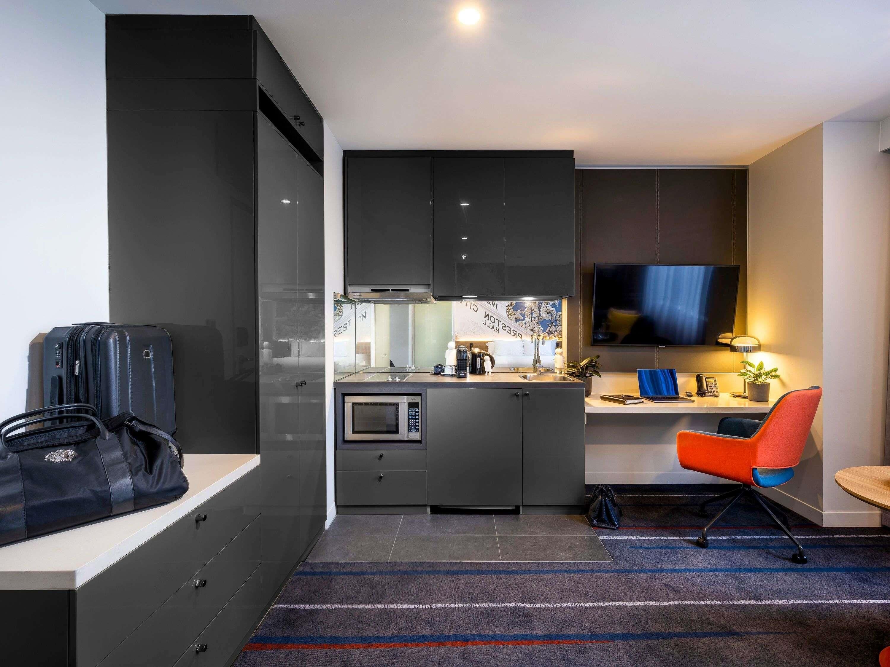 A compact hotel room with a kitchenette, a flat-screen TV, a desk with a chair, and a luggage storage area.