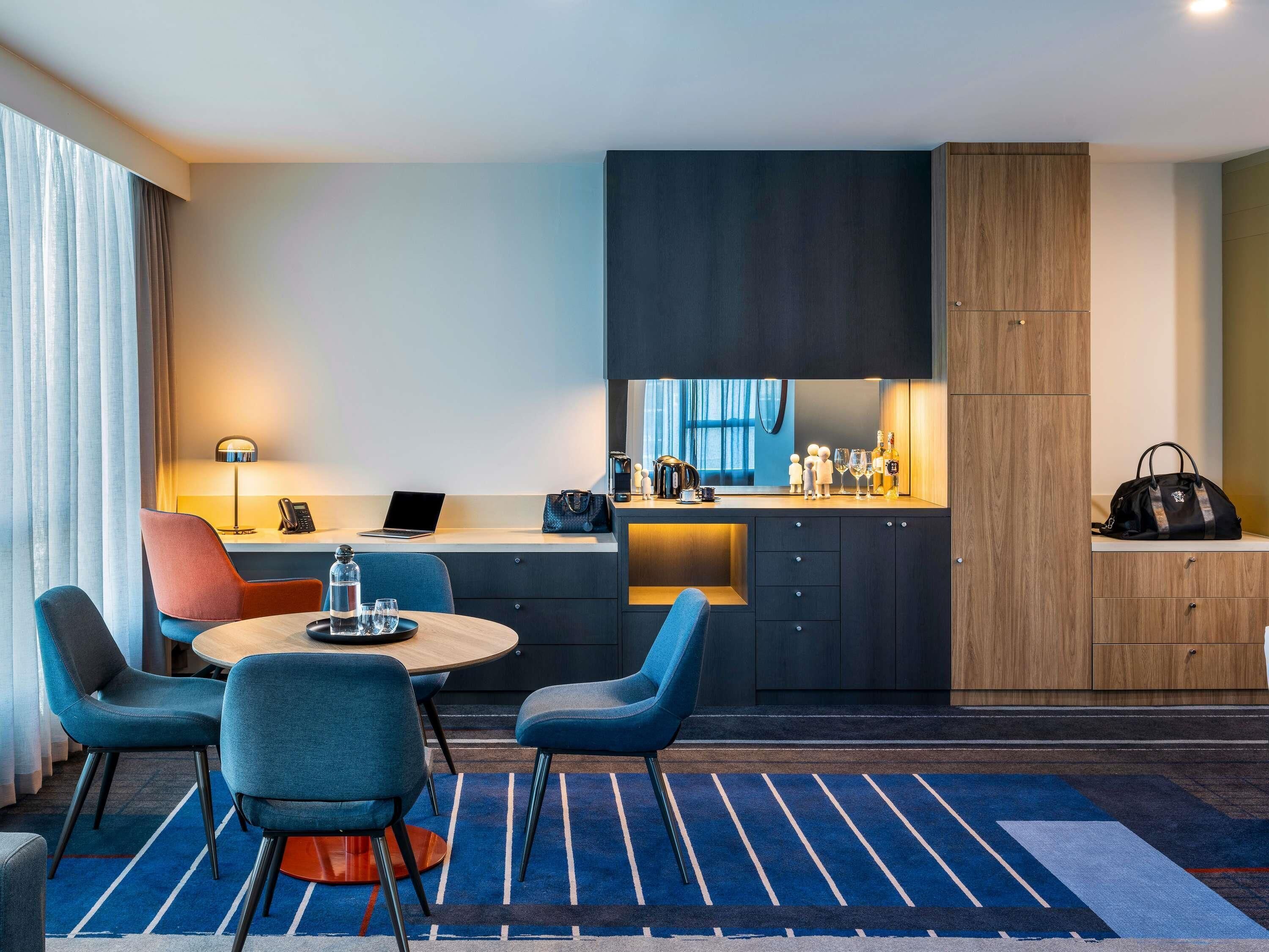 Habitació d'hotel moderna amb zona de menjador, barra de cuina i espai d'estar amb cadires.