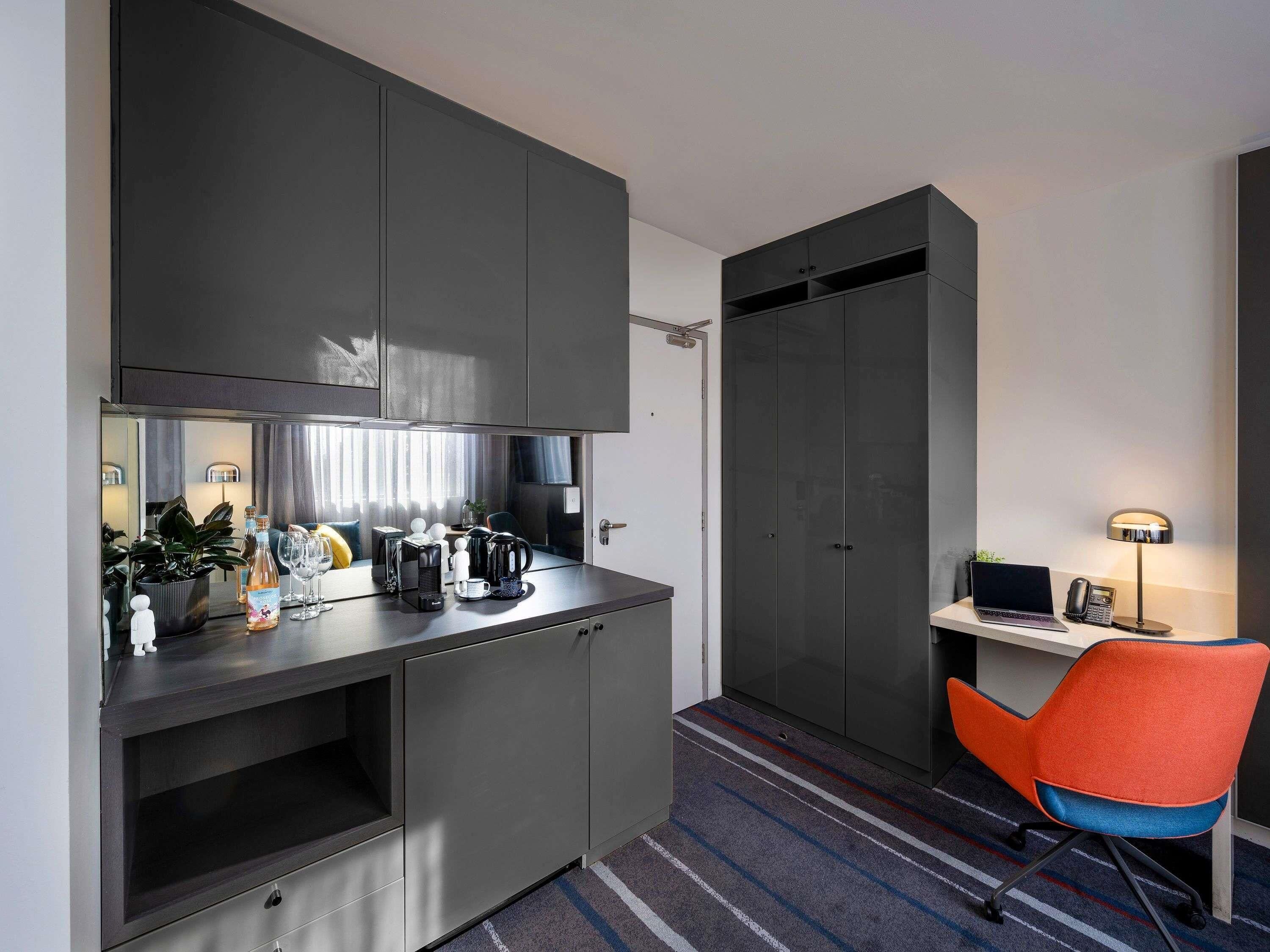 A compact hotel room with a kitchenette, a desk with a laptop and phone, a chair, and a striped carpet.