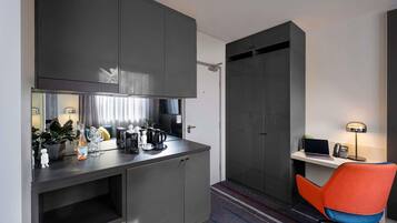 A compact hotel room with a kitchenette, a desk with a laptop and phone, a chair, and a striped carpet.