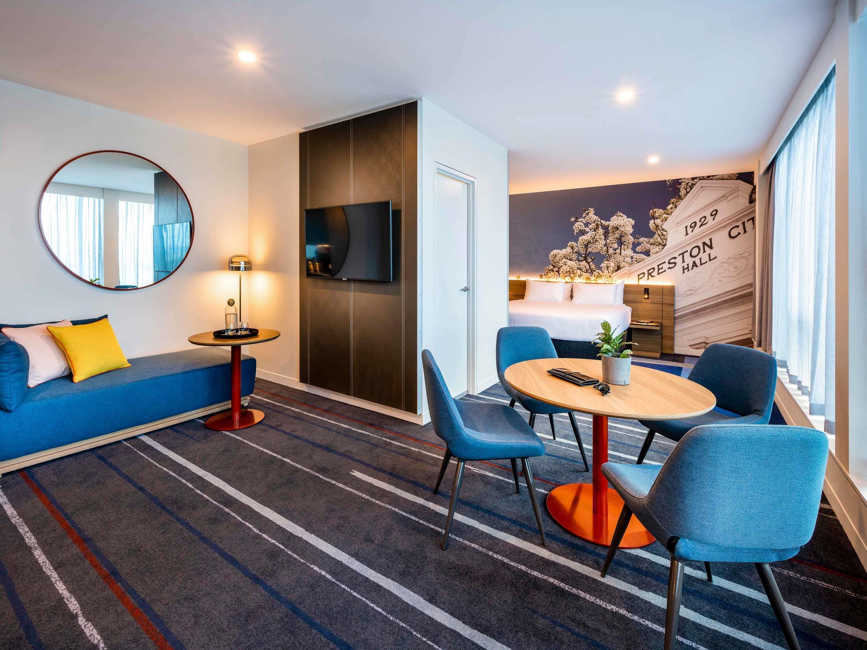 Una habitación de hotel moderna con un sofá, una mesa de comedor redonda con sillas, un televisor y una pared con un mural decorativo.