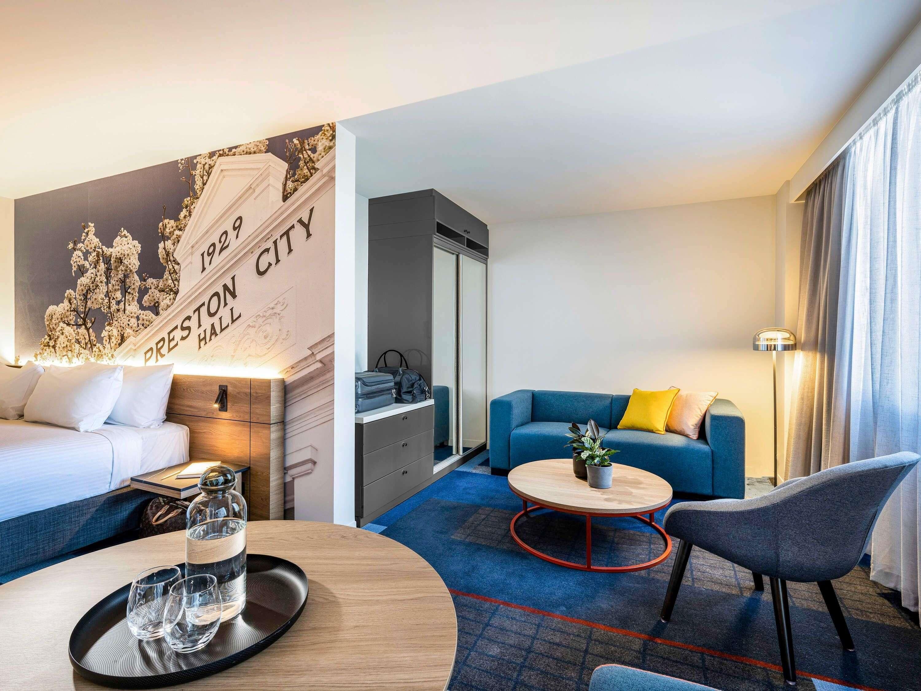 A hotel room with a bed, a sofa, a coffee table, a chair, a desk, a lamp, and a wall with a floral mural.