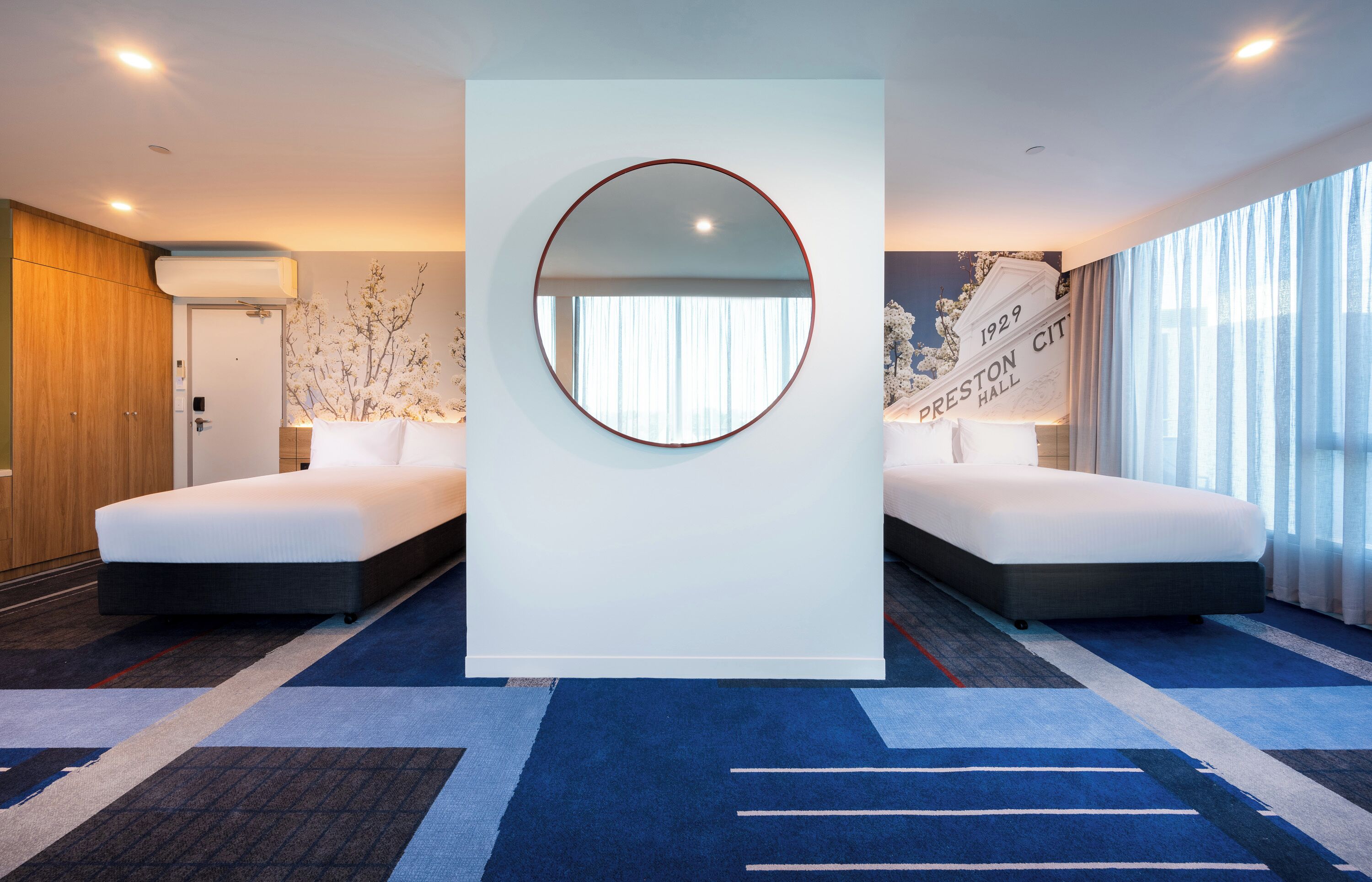 Une chambre d’hôtel avec deux lits, un grand miroir, un climatiseur fixé au mur et une vue sur l’extérieur grâce à de grandes fenêtres.