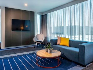 Una habitación de hotel moderna con un sofá, una silla, una mesa de centro redonda de madera, un televisor montado en la pared y ventanas amplias con cortinas traslúcidas.