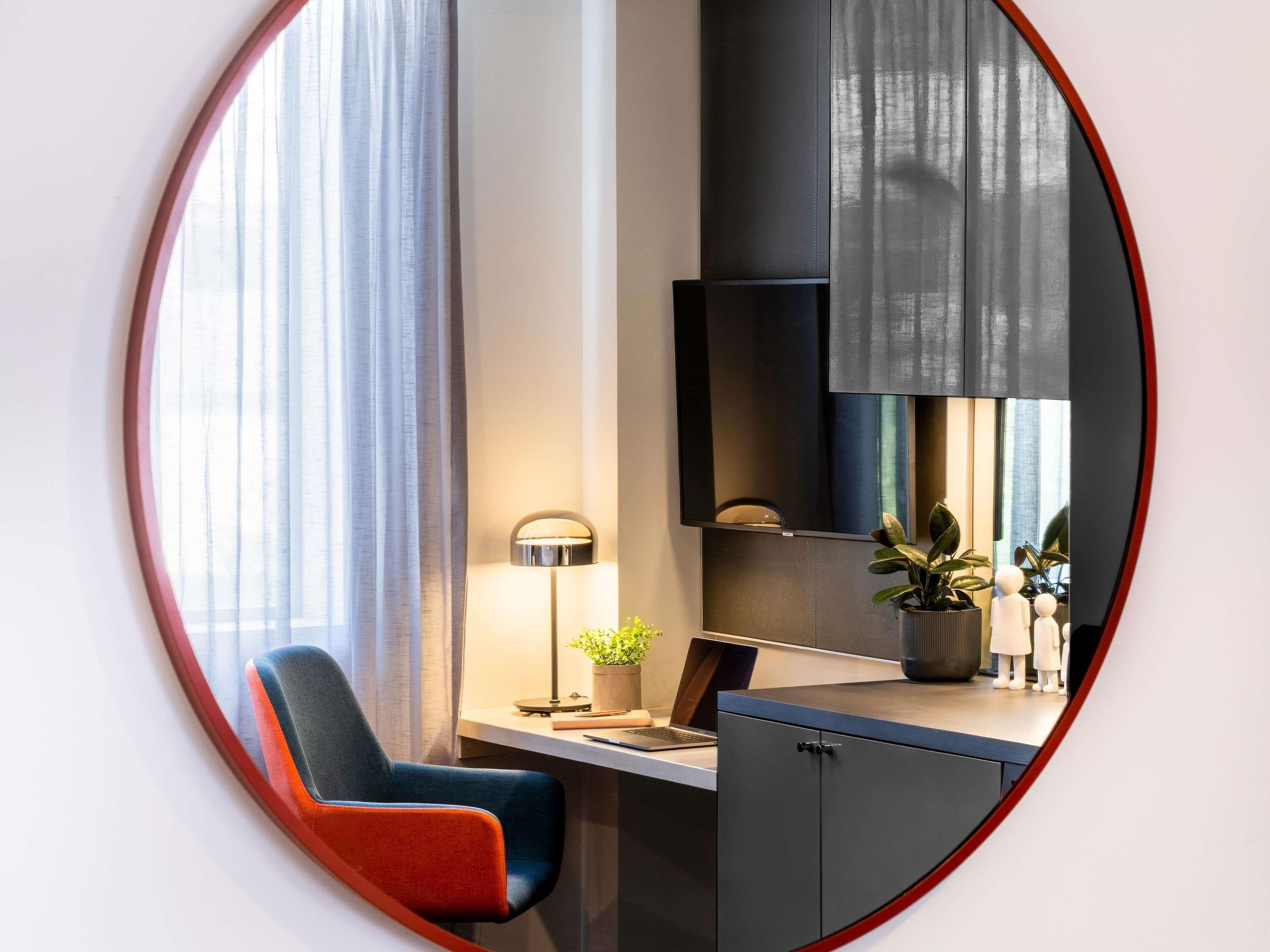 Una habitación de hotel moderna con escritorio, silla, lámpara, planta en maceta y un espejo grande.