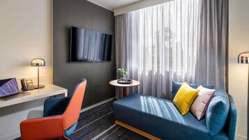 Habitación de hotel con sofá, escritorio, laptop, silla, televisor y ventana con cortinas.