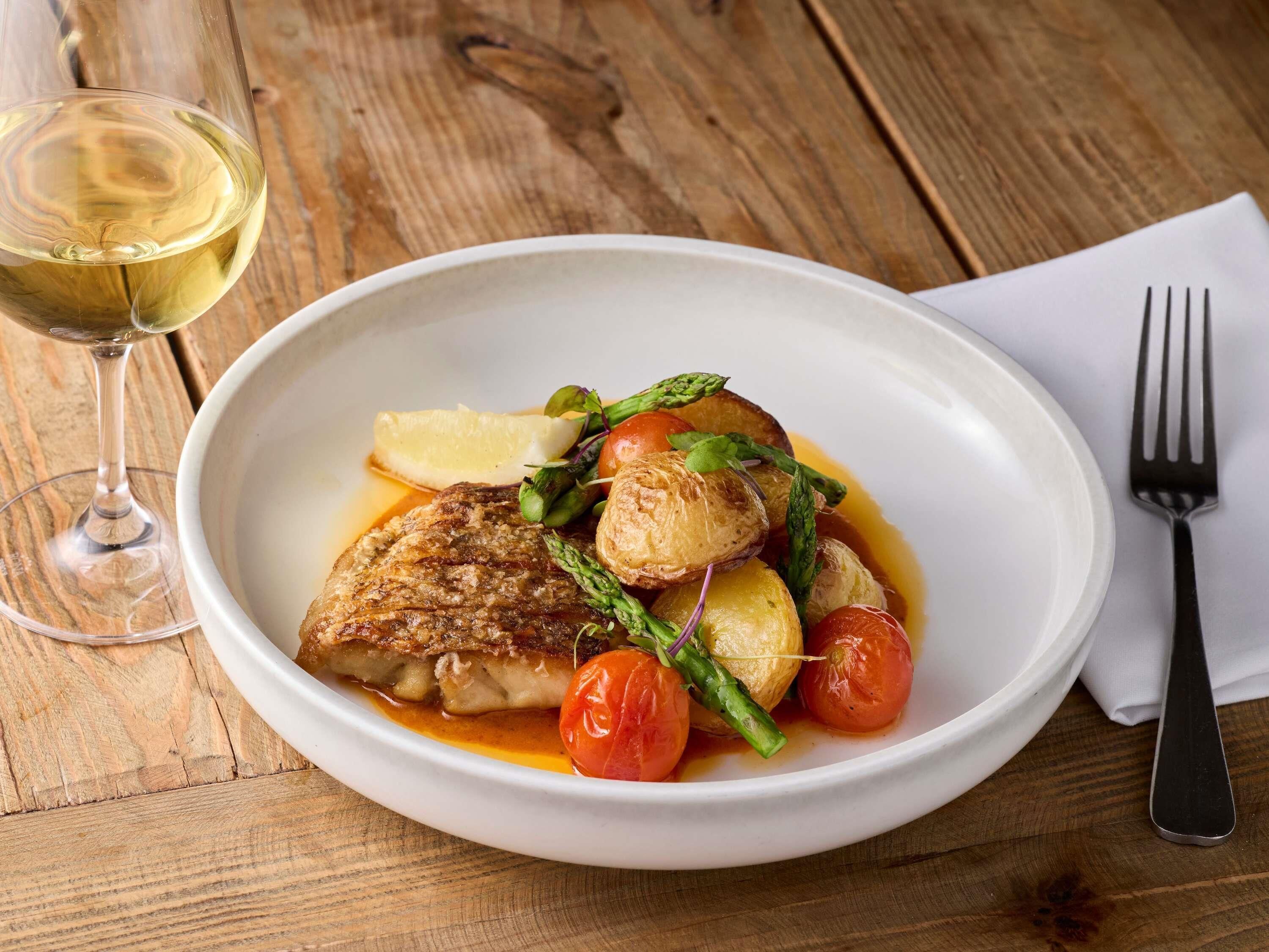 A plate of food with a fish, asparagus, and tomatoes, accompanied by a glass of white wine on a wooden table.