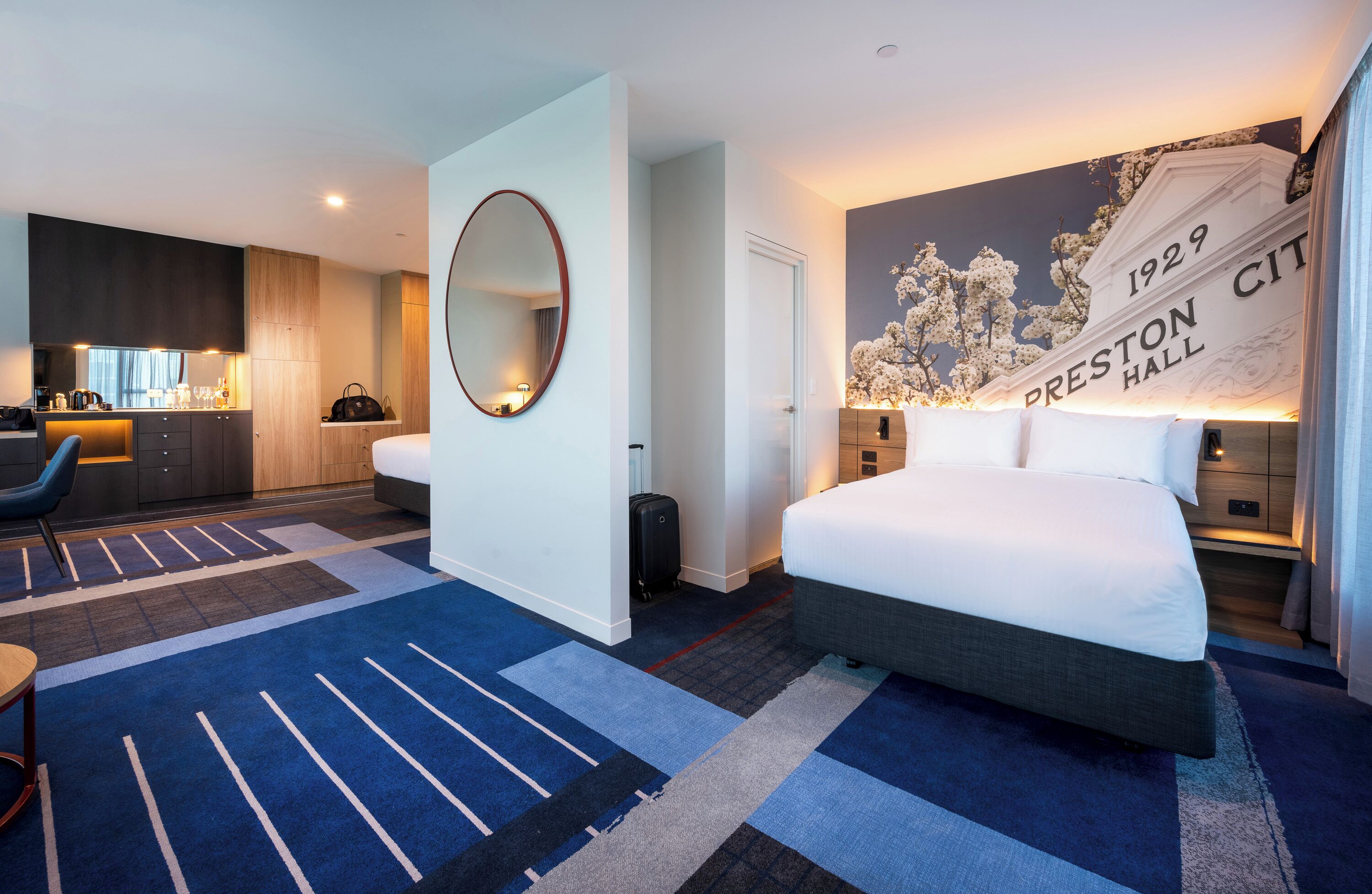 A hotel room with a large bed, a desk with a chair, a kitchenette, a large mirror, and a wall with a plaque reading "1929 Preston Hall".