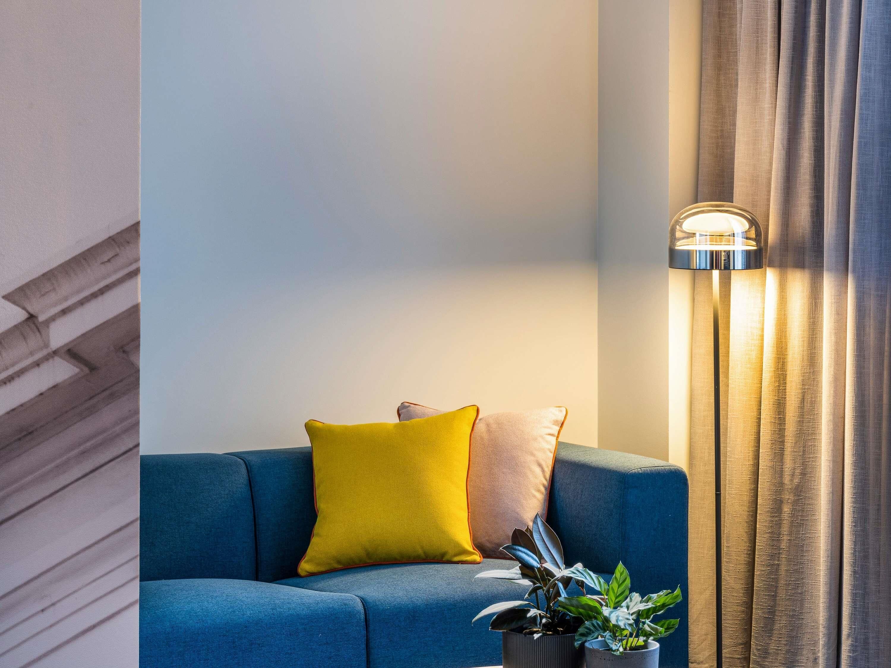 Um sofá azul com almofadas amarelas e castanhas, uma luminária de chão e um vaso com planta.