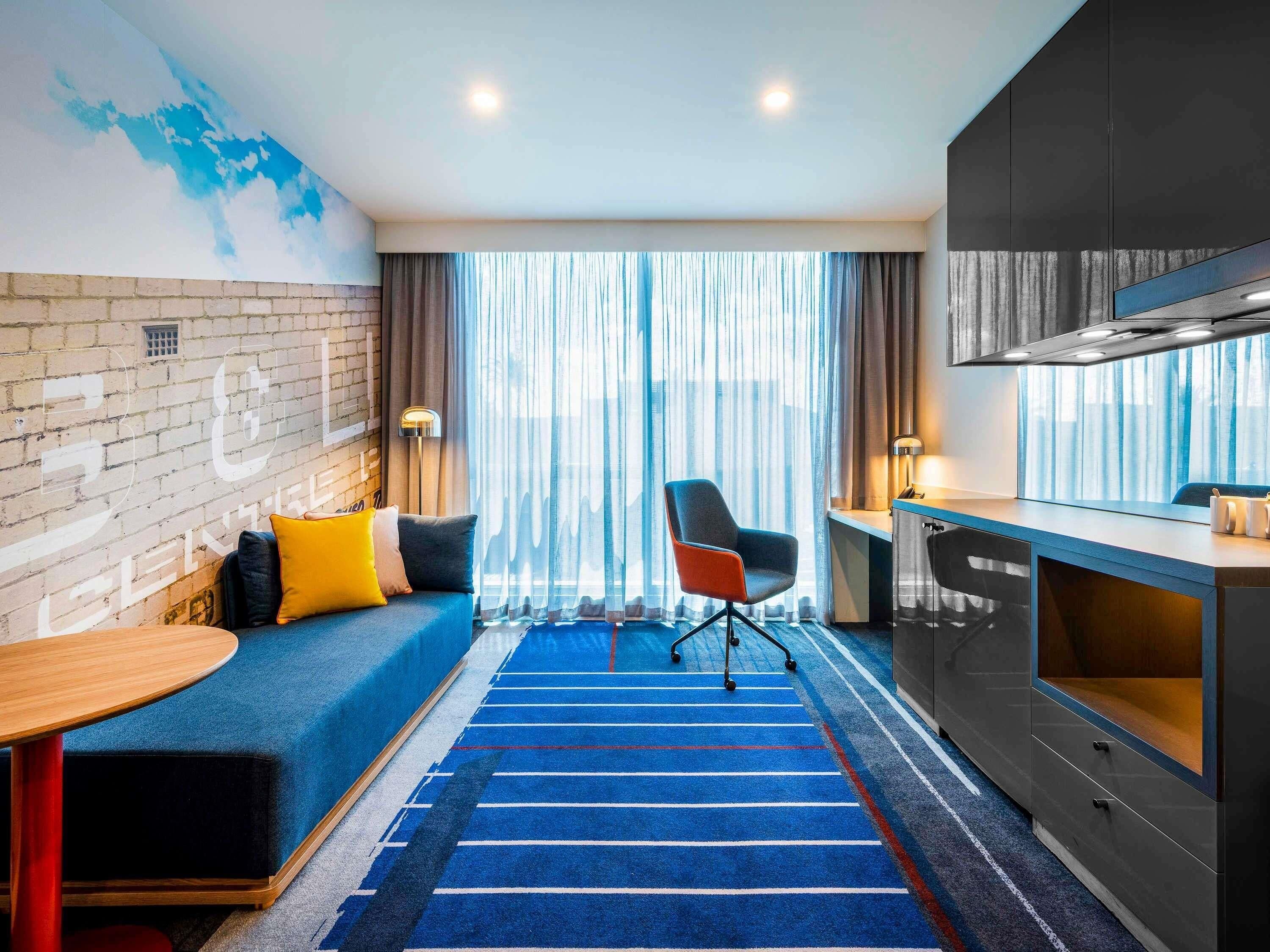 Ein modernes Hotelzimmer mit blauem Teppich, einer Couch mit Kissen, einem Schreibtisch mit Lampe und einer Küchenzeile.