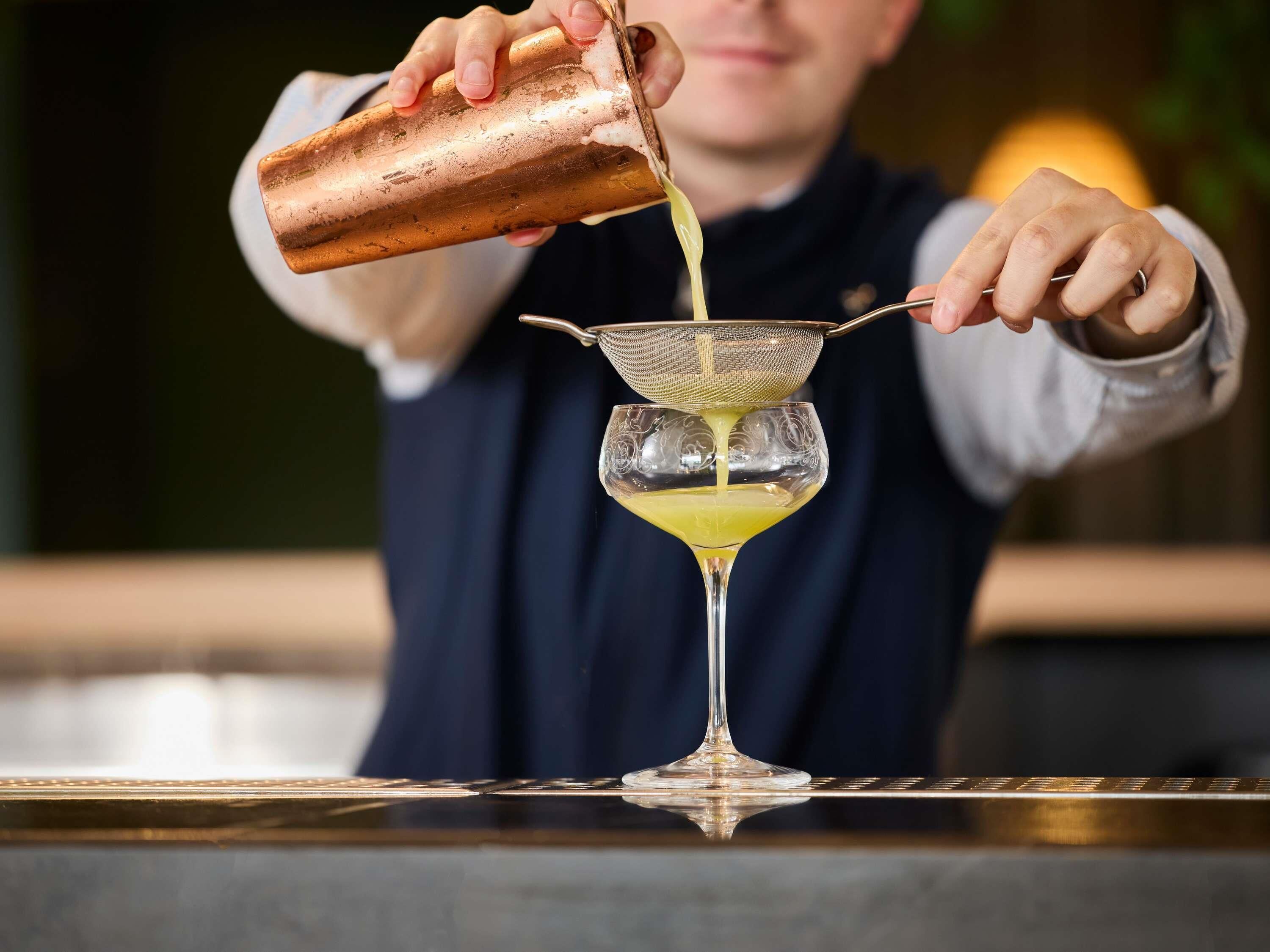 A bartender pouring a yellow liquid into a martini glass using a strainer.