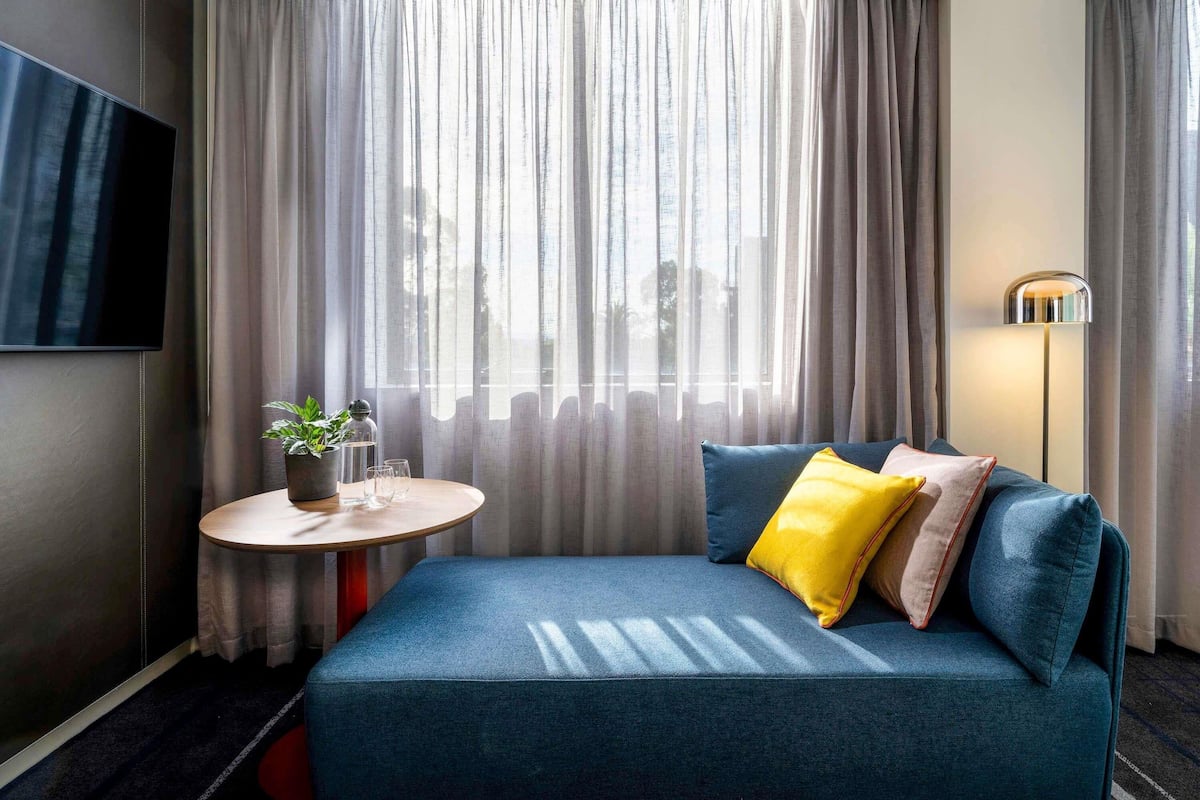 Una habitación de hotel moderna con un sofá azul, una mesita redonda con una planta, un televisor de pantalla plana y una lámpara de pie.