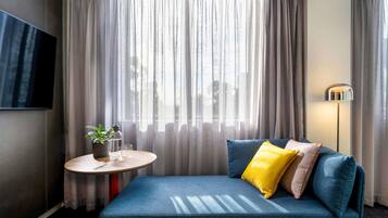 Una habitación de hotel moderna con un sofá azul, una mesita redonda con una planta, un televisor de pantalla plana y una lámpara de pie.