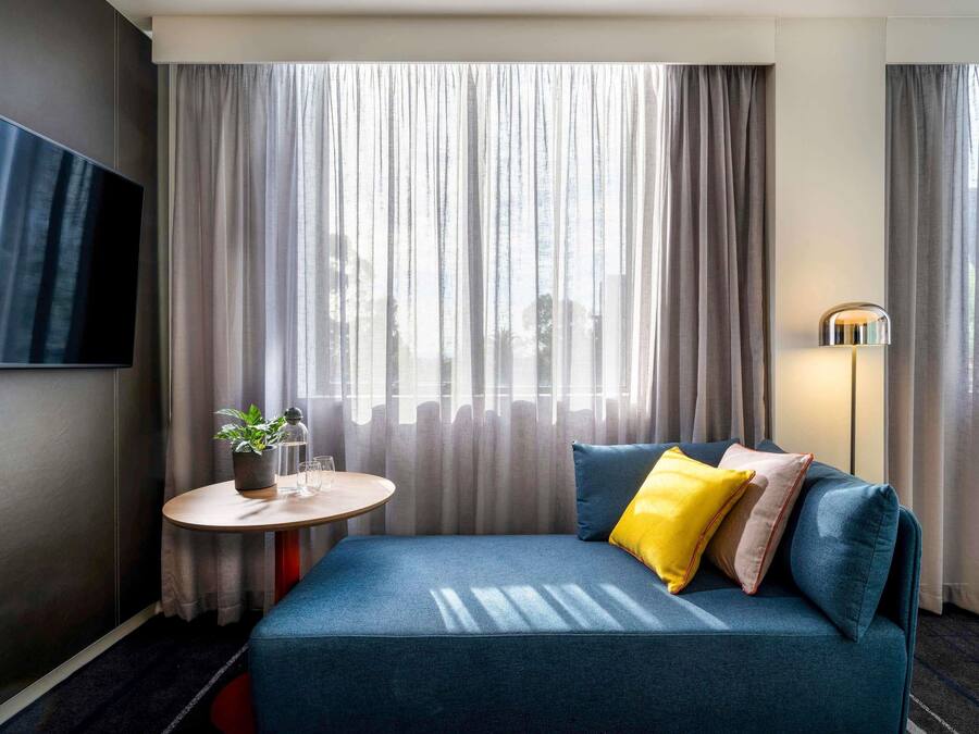 A modern hotel room with a blue sofa, a small round table with a plant, a flat-screen TV, and a floor lamp.