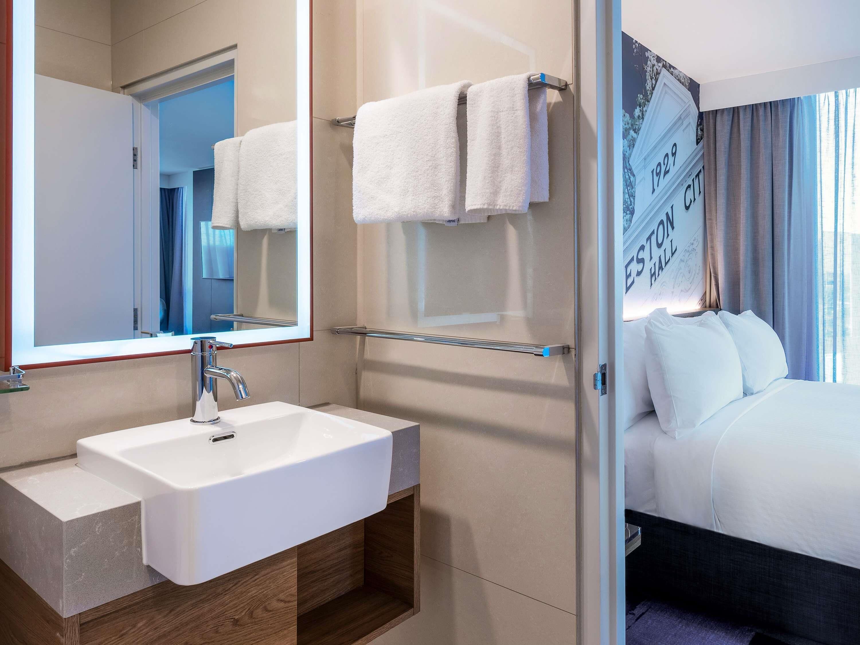 A hotel room with a large mirror, a white sink, a glass shower enclosure, and a bed with white bedding.